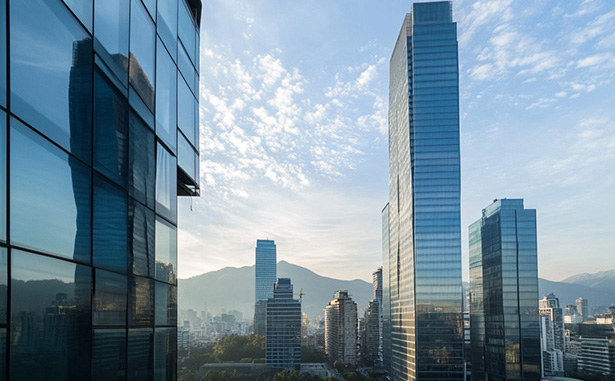Vista de una ciudad con varios rascacielos modernos de superficies reflectantes. El cielo está parcialmente nublado con un tono azul y, en el fondo, se pueden ver montañas bajo el cielo despejado.