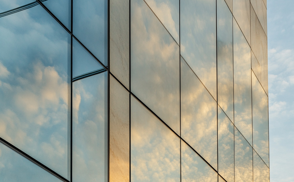 Edificio moderno con fachada de vidrio reflectante que refleja el cielo parcialmente nublado, creando un efecto visual en el que las nubes parecen estar integradas en la estructura del edificio.