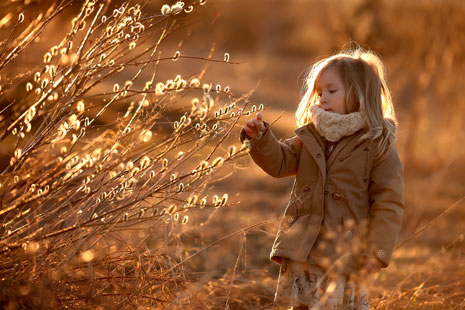 Niña con abrigo color café al aire libre, rodeado de plantas con tonos dorados. La niña está extendiendo la mano hacia las plantas, que están iluminadas por la cálida luz del sol.